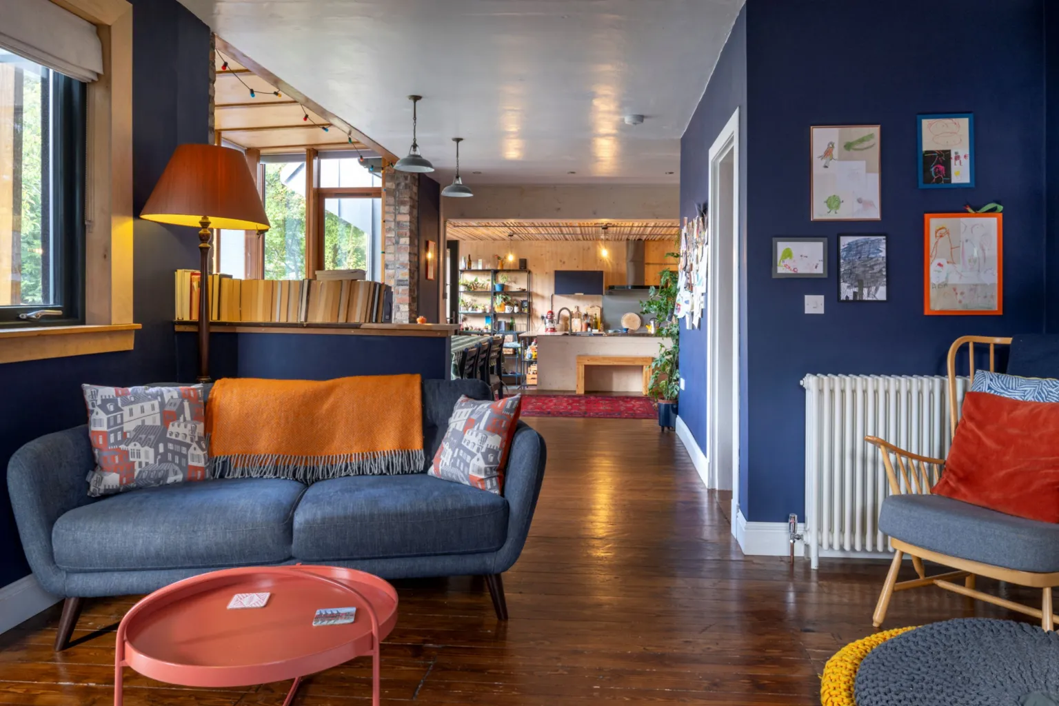 IWC Media A different view of the living area shows more of the warm orange touches, a lamp, a velvet cushion on a wooden crafted chair, warm lights in the kitchen beyond. Children's paintings are framed on the wall.