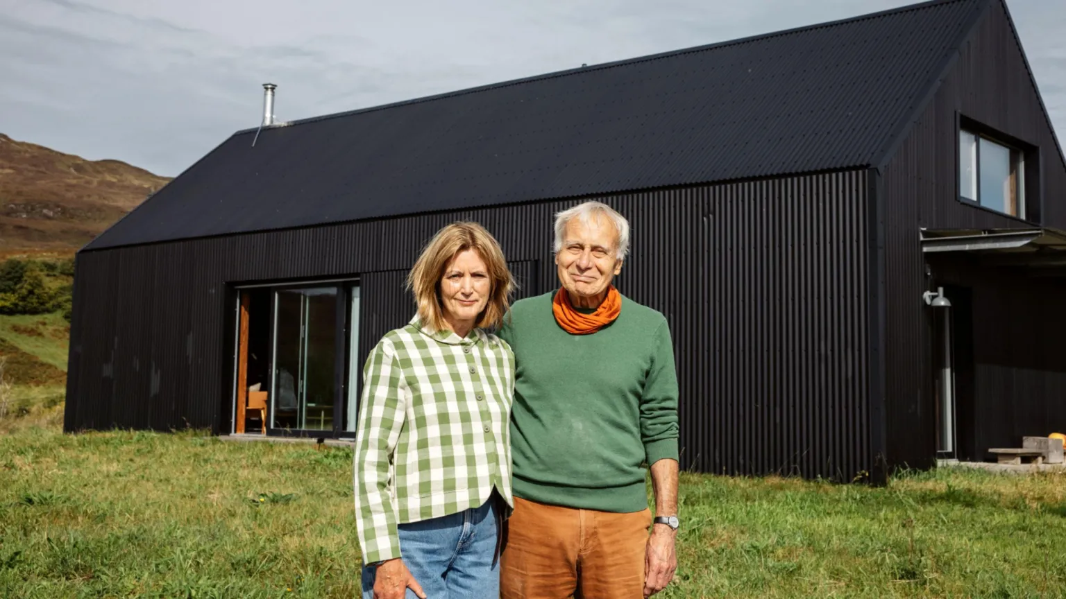 IWC Media Two people stand side by side in a grassy outdoor area in front of a modern, dark-clad house with a sloped roof. The building features large windows, a metal chimney pipe, and a minimalist design set against a rural hillside landscape. The scene is lit by natural daylight, with muted skies and earthy tones highlighting the contrast between the black structure and the surrounding greenery.