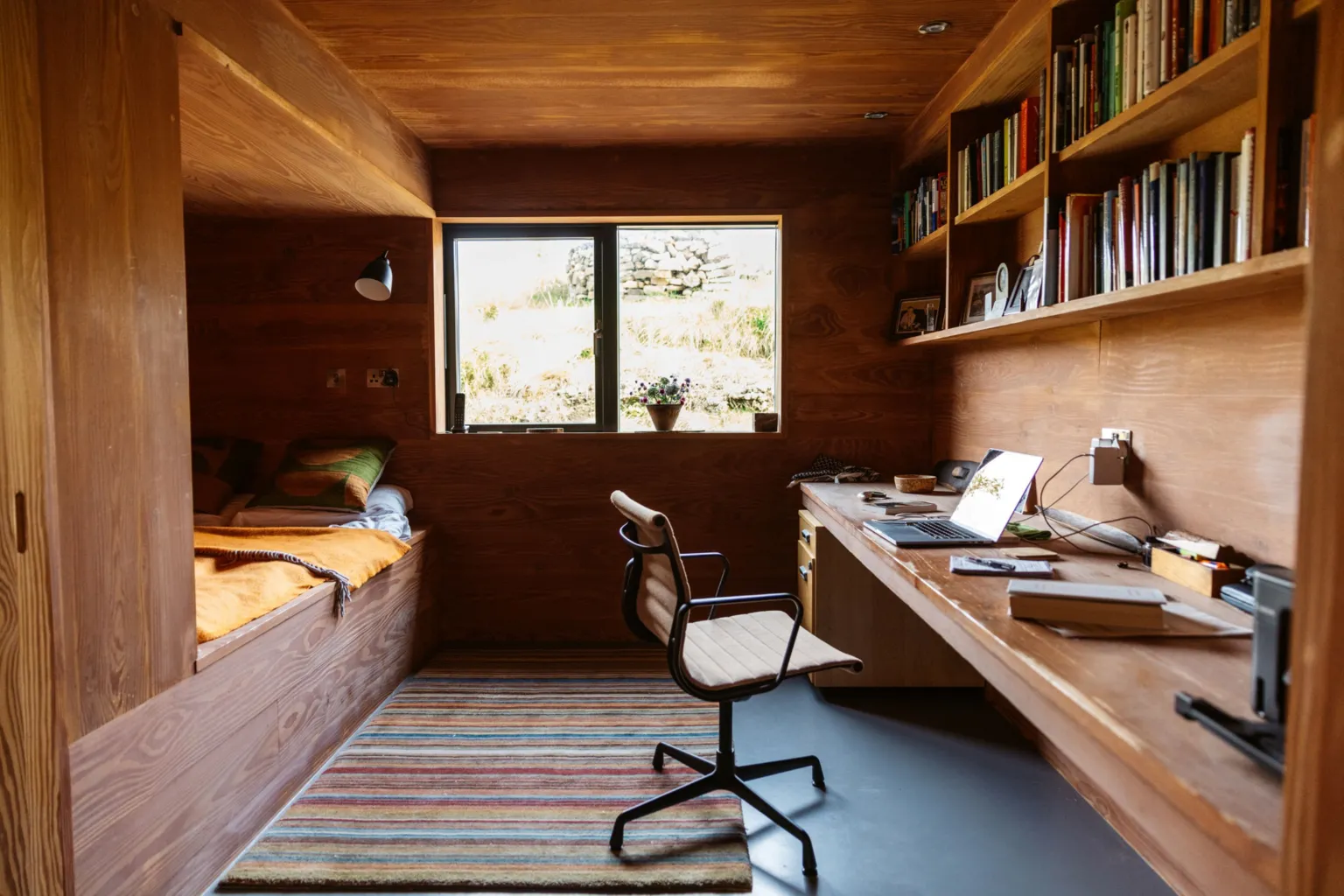 IWC Media A compact, wood-panelled room features a built-in bed on the left with cushions and a mustard-coloured blanket. Opposite, a long wooden desk holds an open laptop, books, and small objects, with shelves above filled with neatly arranged books and framed photos. A window centred on the back wall lets in daylight and reveals a grassy outdoor scene, while a striped rug and simple chair complete the cosy workspace.