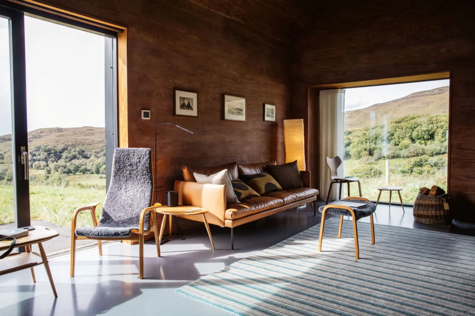 IWC Media A warm, wood-panelled living room features a leather sofa, minimalist chairs, and a striped rug arranged in a clean, modern layout. Large floor-to-ceiling windows frame a sunlit rural landscape, blending the cosy interior with the surrounding greenery.