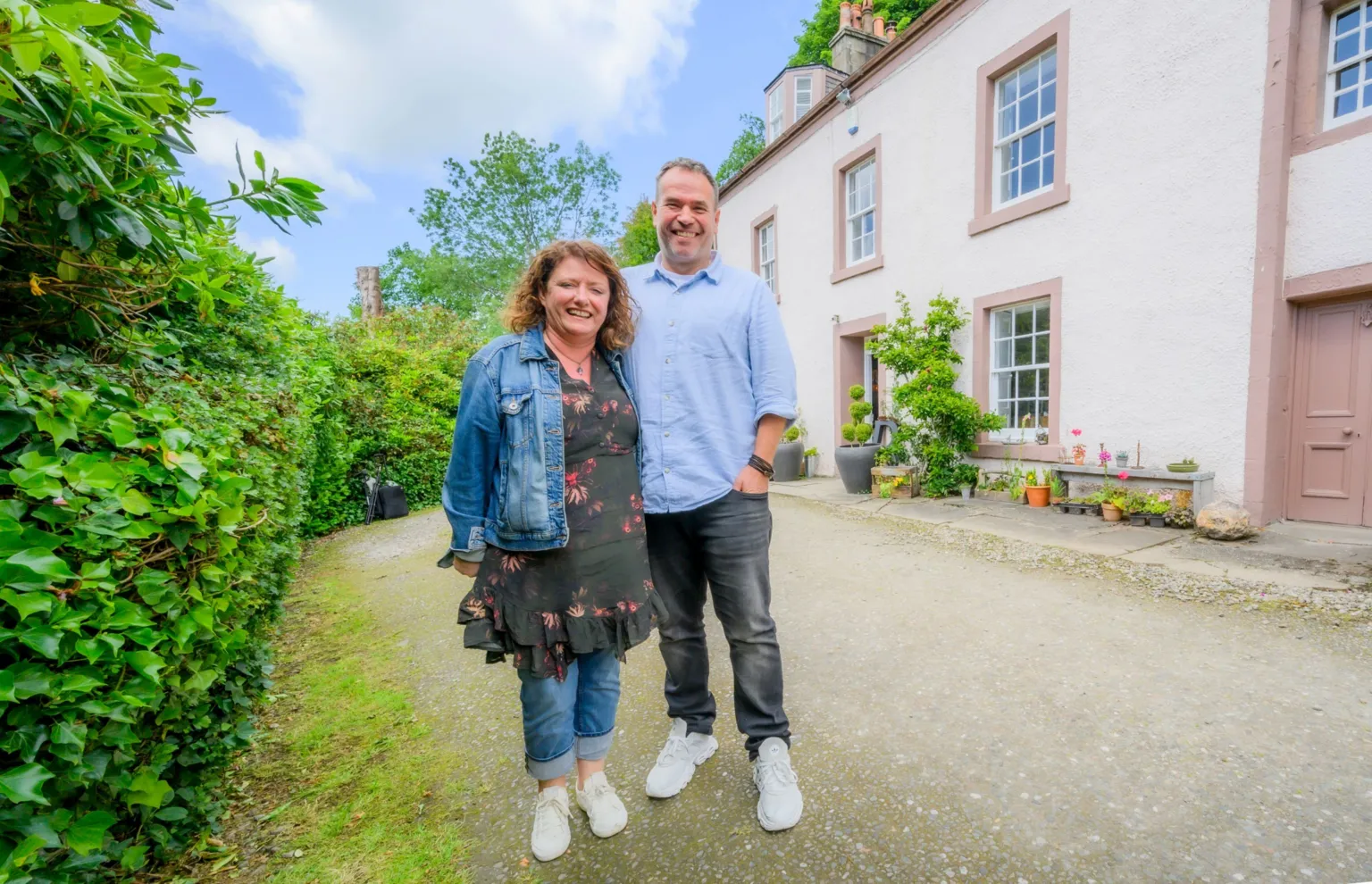 IWC Media Linda and Euan stand side by side on a gravel driveway in front of a light-coloured house with large windows and a doorway. Lush green hedges line the left side, while potted plants and small garden features decorate the area near the building. The scene is brightly lit under a partly cloudy sky, giving a vibrant and airy outdoor setting.