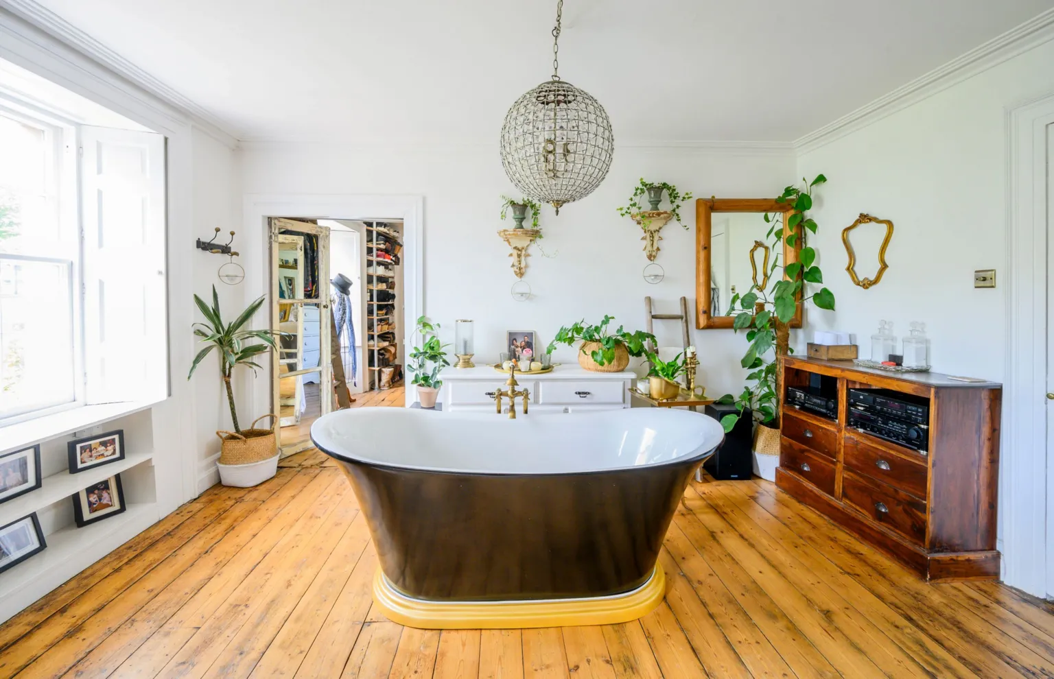 IWC Media A bright, spacious bathroom features a freestanding black bathtub placed centrally on warm wooden floorboards. White walls are decorated with mirrors, plants, and shelves, while a large window lets in abundant natural light. A chandelier hangs above, and vintage furniture and accessories add a stylish, eclectic touch to the room.