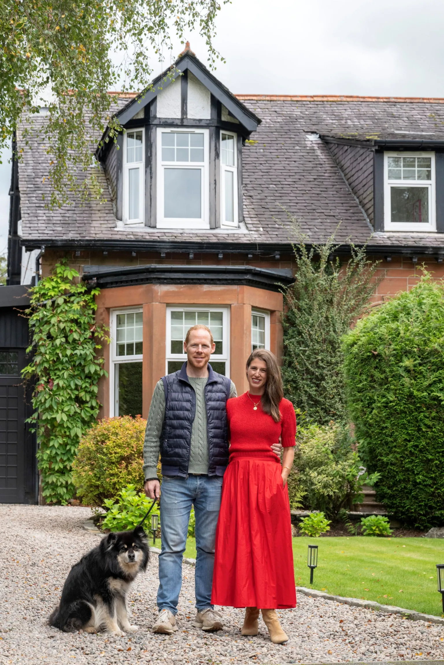 IWC Media Scott and Wendy stand side by side on a gravel driveway beside a black dog, in front of a large house with a steep, tiled roof and dormer windows. The house features warm stone walls, black timber accents, and is surrounded by neatly maintained greenery and shrubs. Overhanging tree branches and soft daylight create a calm, residential atmosphere in the garden setting.