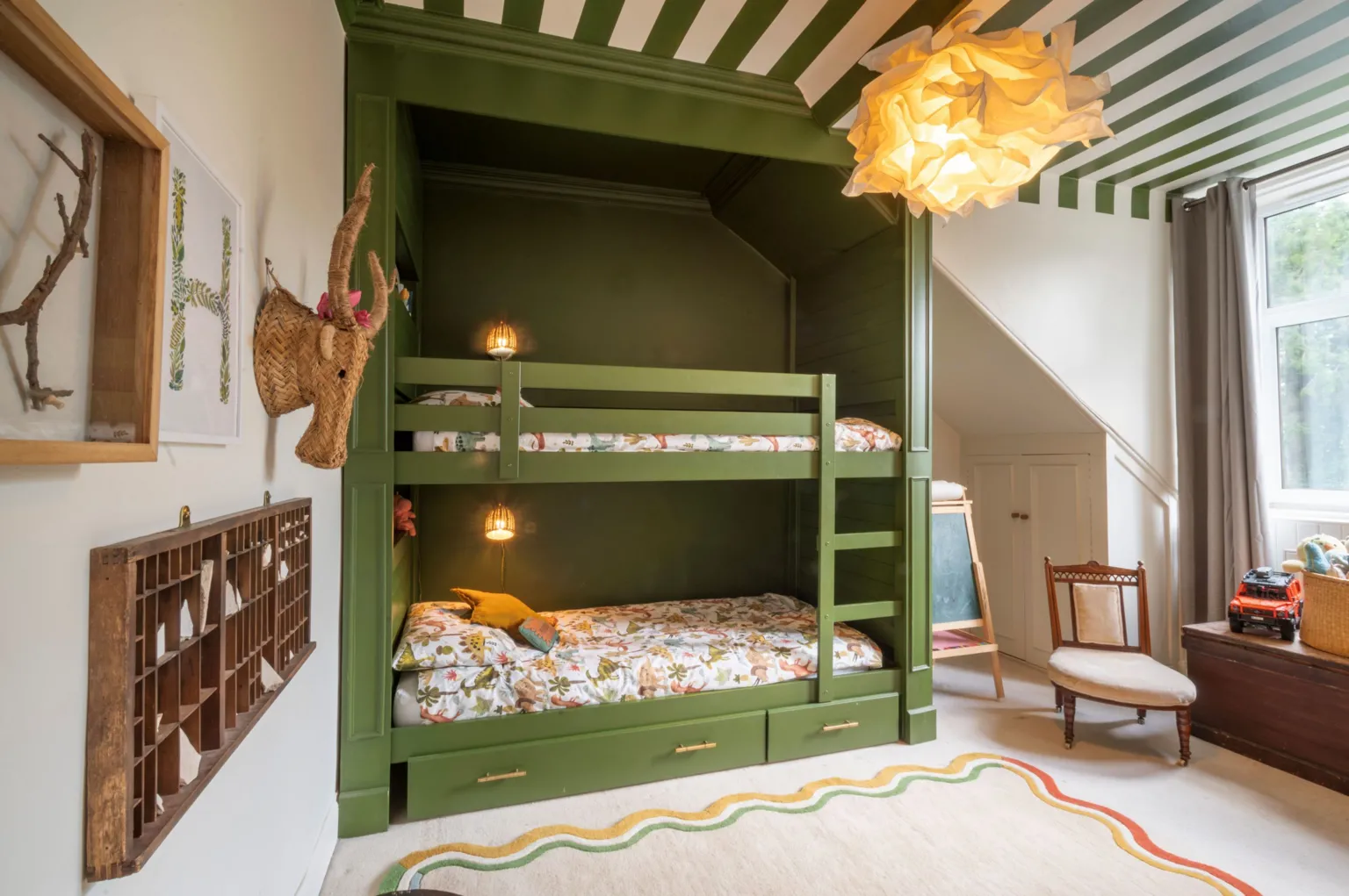 IWC Media A cosy children’s bedroom features a green built-in bunk bed with patterned bedding and warm wall lights tucked into the frame. Playful decor, including a woven animal head, framed artwork, toys, and a soft rug, creates a bright and inviting space near a large window.
