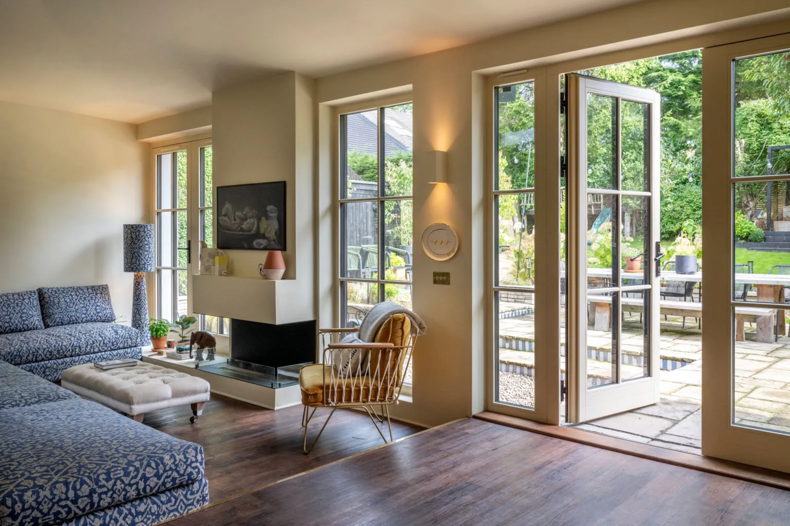 IWC Media A bright, modern living room features large glass doors and windows that open onto a green garden with a patio and outdoor seating. Inside, a blue patterned sofa, neutral ottoman, and a sleek fireplace create a comfortable seating area with soft, natural lighting.
