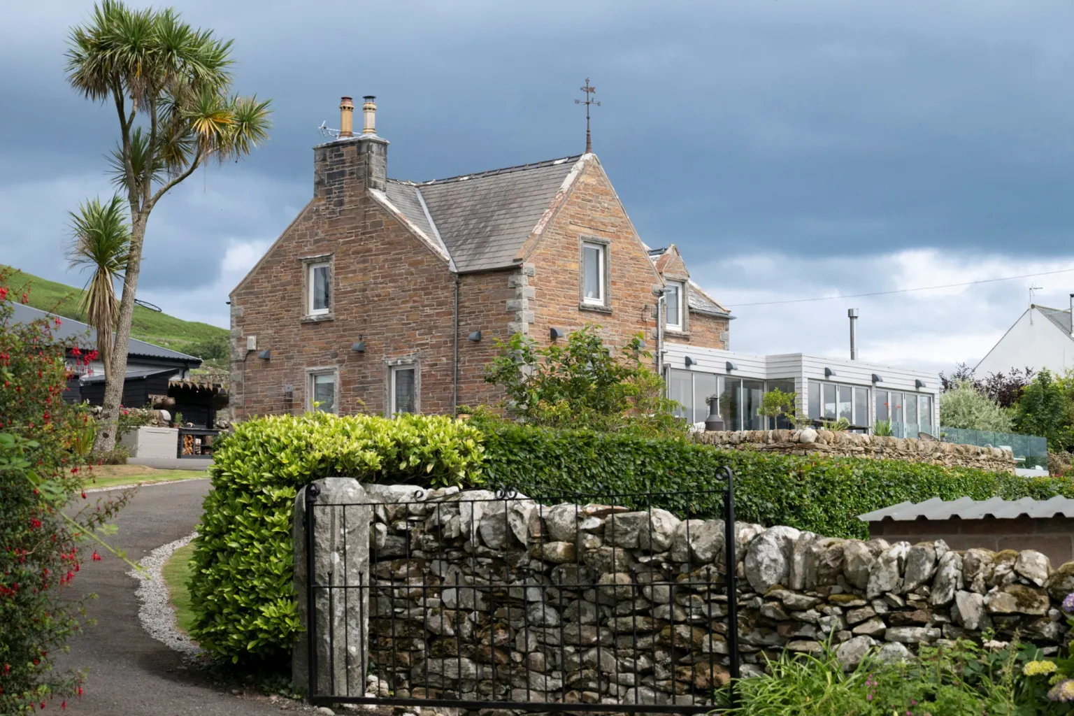 IWC Media A traditional stone house with a pitched roof sits behind a low stone wall and iron gate, surrounded by lush green shrubs and garden plants. A tall, spiky tree stands to the left, while a modern extension with large windows is visible on the right side of the building. Dark, heavy clouds fill the sky above, contrasting with the warm tones of the stone exterior and the greenery below.