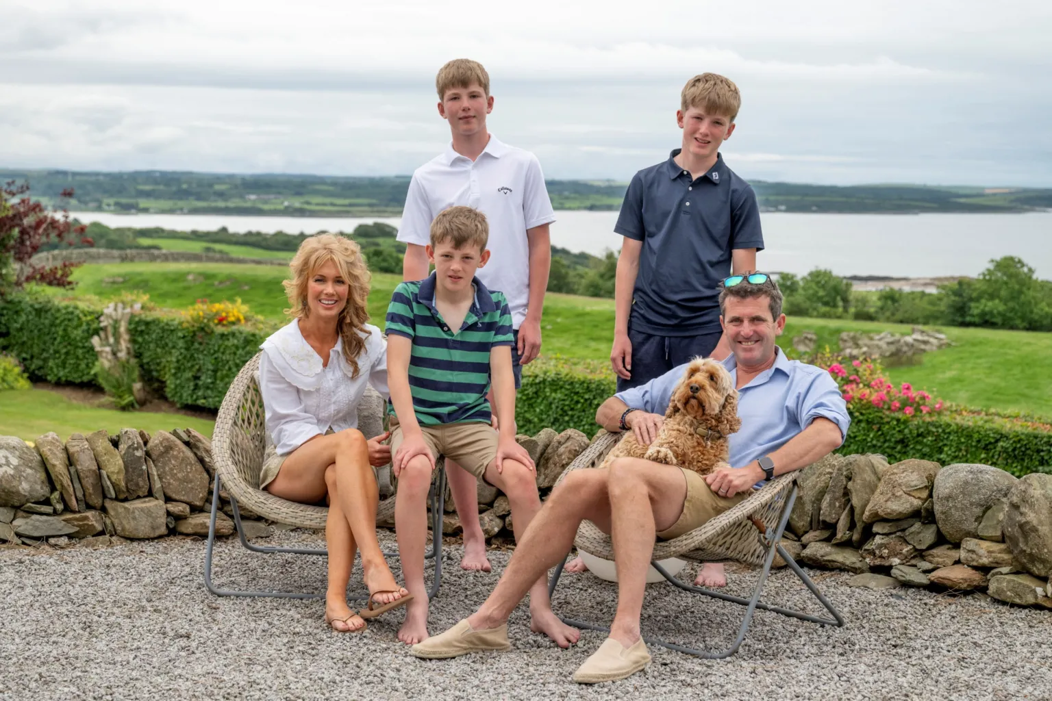 IWC Media A group of five people sit and stand together outdoors on gravel beside a low stone wall, accompanied by a small dog resting on a seated person’s lap. Behind them stretches a lush green landscape with rolling fields and a calm body of water under a cloudy sky. Casual seating, light clothing, and garden flowers create a relaxed, scenic setting in the countryside.