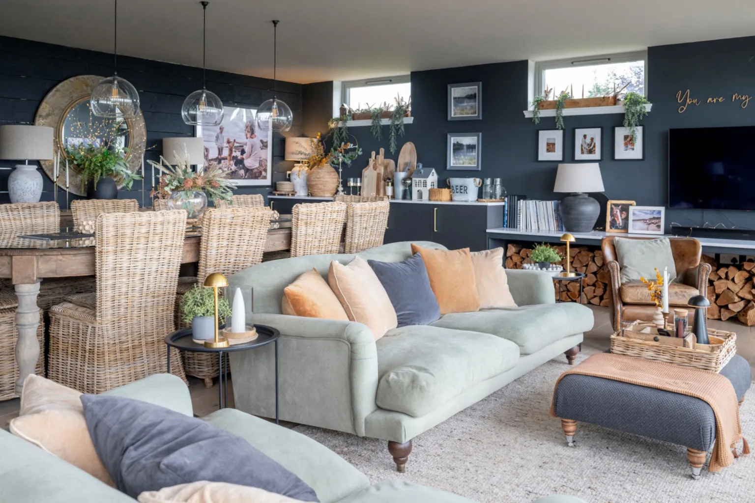 IWC Media A stylish open-plan living and dining area features soft green sofas, woven chairs, and layered neutral decor against dark painted walls. Glass pendant lights hang above a wooden dining table, while shelves, plants, and framed photos add warmth and personality. 