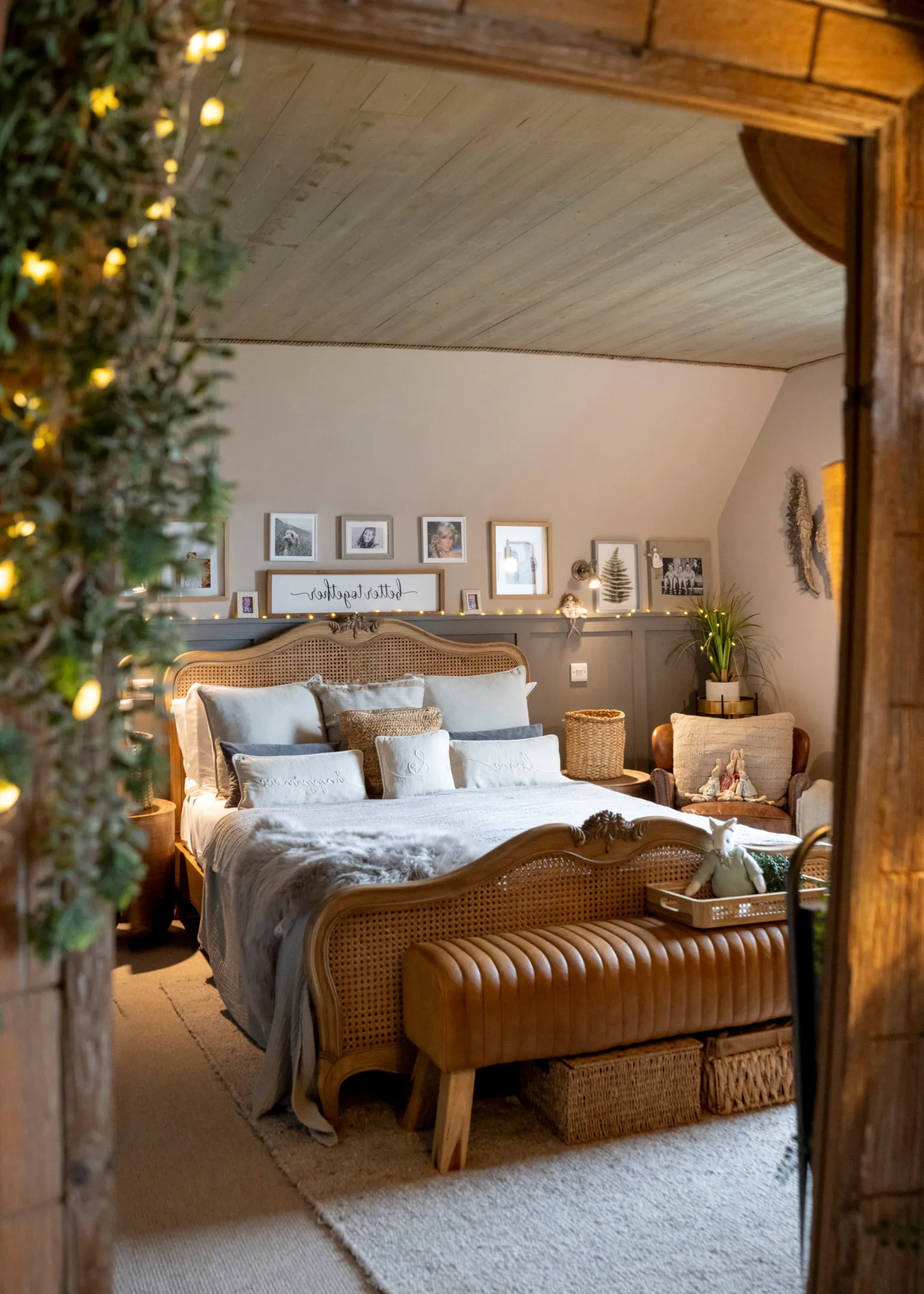 IWC Media A warm, cosy bedroom features a wooden-framed bed layered with soft pillows and neutral-toned bedding beneath a sloped ceiling. Framed photos, plants, and string lights decorate the walls, while a cushioned bench and woven baskets add texture and comfort.