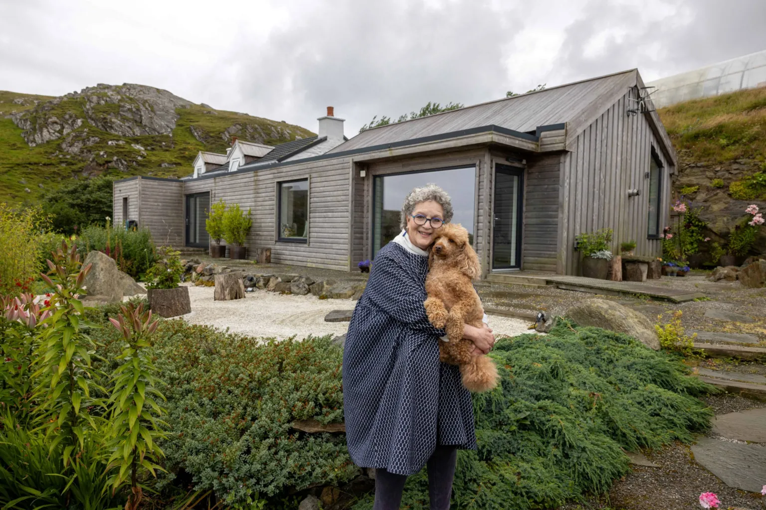 IWC Media Susie stands in a landscaped garden holding a small dog, in front of a modern, wood-clad house set against rolling hills. The home features large windows, clean lines, and a mix of rustic and contemporary design elements. Surrounding plants, stone paths, and a cloudy sky create a calm, natural countryside setting.