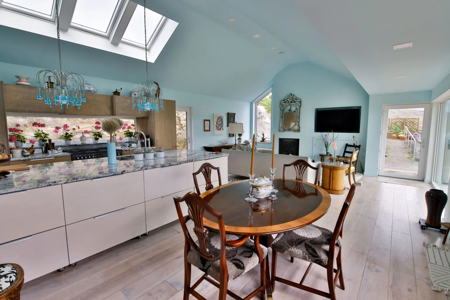 IWC Media A bright, open-plan kitchen and dining space features pale blue walls, skylights, and large windows that flood the room with natural light. A marble-topped island, modern cabinetry, and a round wooden dining table with chairs create a blend of contemporary and classic design.