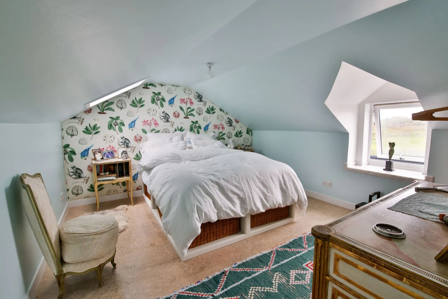 IWC Media A bright attic bedroom features a low, sloping ceiling painted pale blue and a bed dressed in crisp white bedding. A playful patterned wall with plants and animals, along with small furniture and a window, adds charm and character to the cosy space.