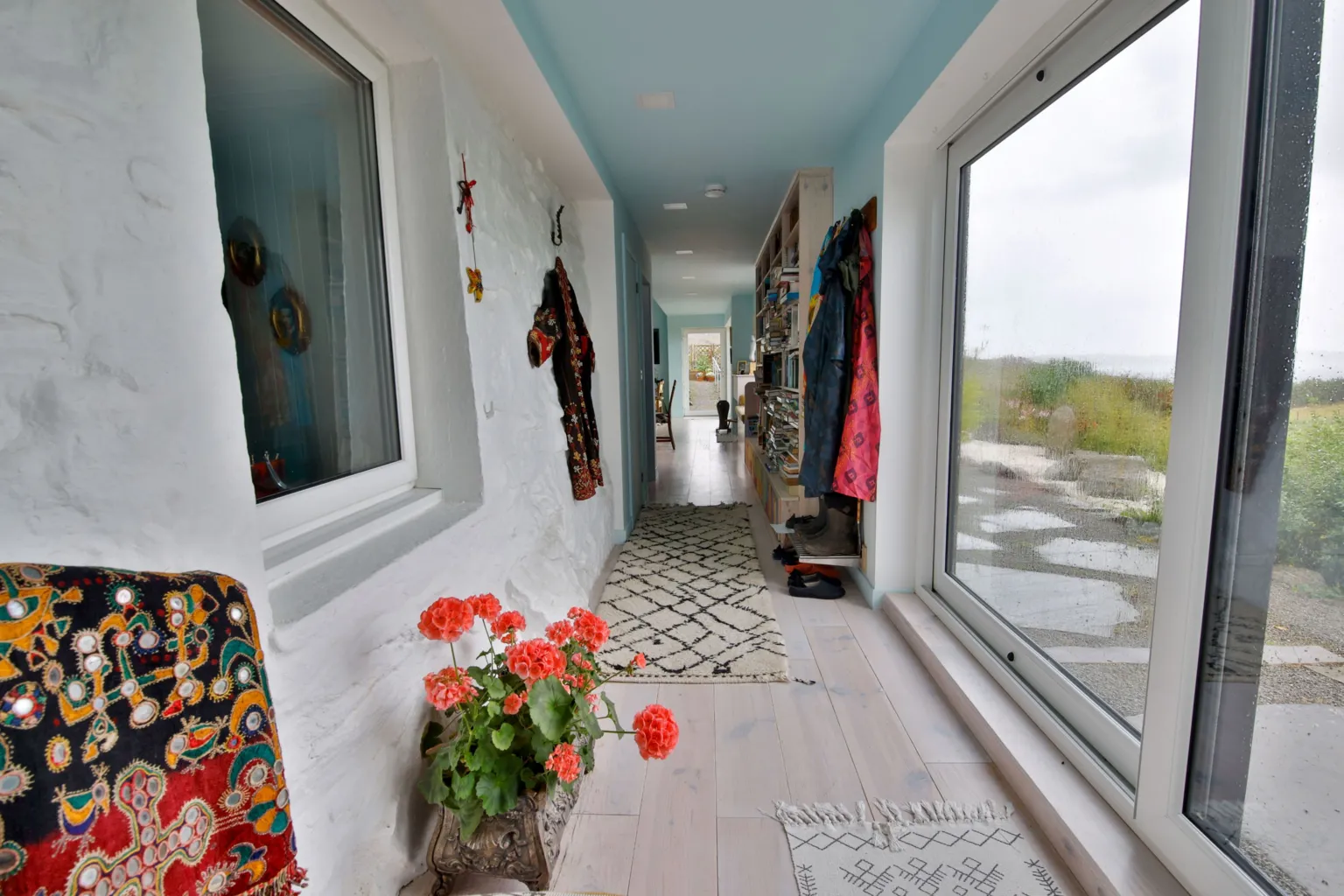 IWC Media A narrow, light-filled hallway features textured white walls, pale wood flooring, and a ceiling painted soft blue. Large sliding glass doors line one side, while coats, bookshelves, and a patterned rug add colour and lived-in detail along the corridor.