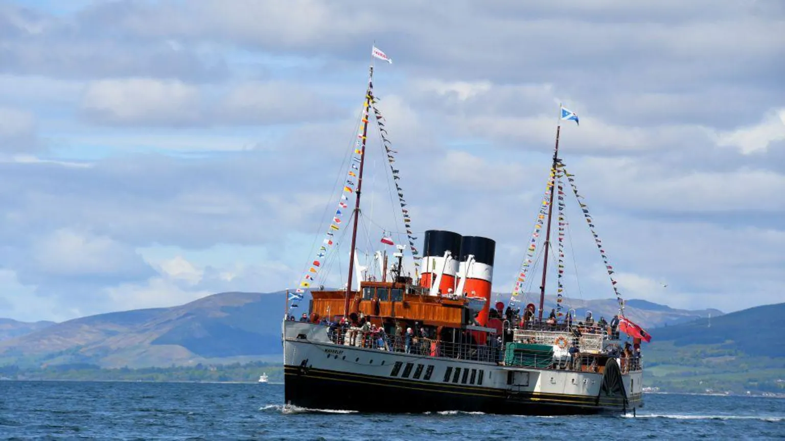 Sylvia Watson The boat 'Waverley' sailing through water, with its famous black, white and red funnels, and bunting and flags, with hills in the distance, under a cloudy blue sky.
