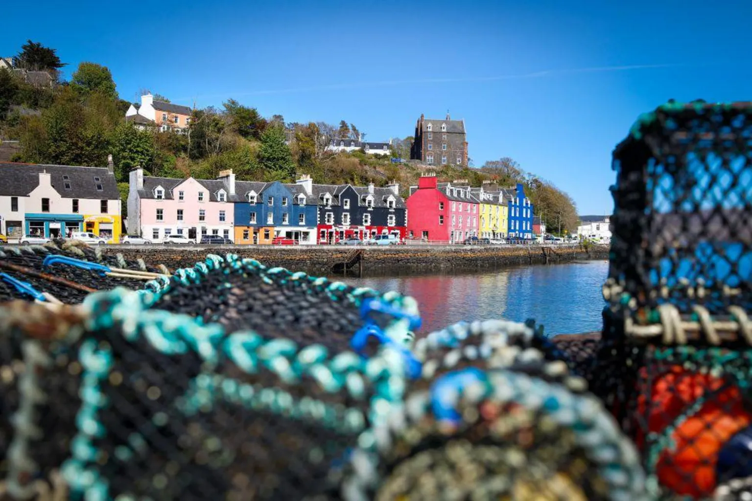 Alex Mackintosh Colourful buildings - including blue, yellow and pink - on a bright sunny day, with lobster pots out of focus in the foreground.