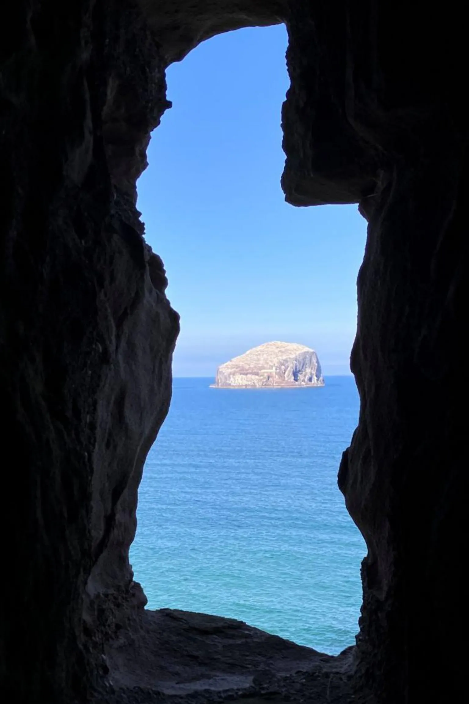 Diane Williamson Bass Rock, a majestic island in the sea, photographed through a dark craggy frame.