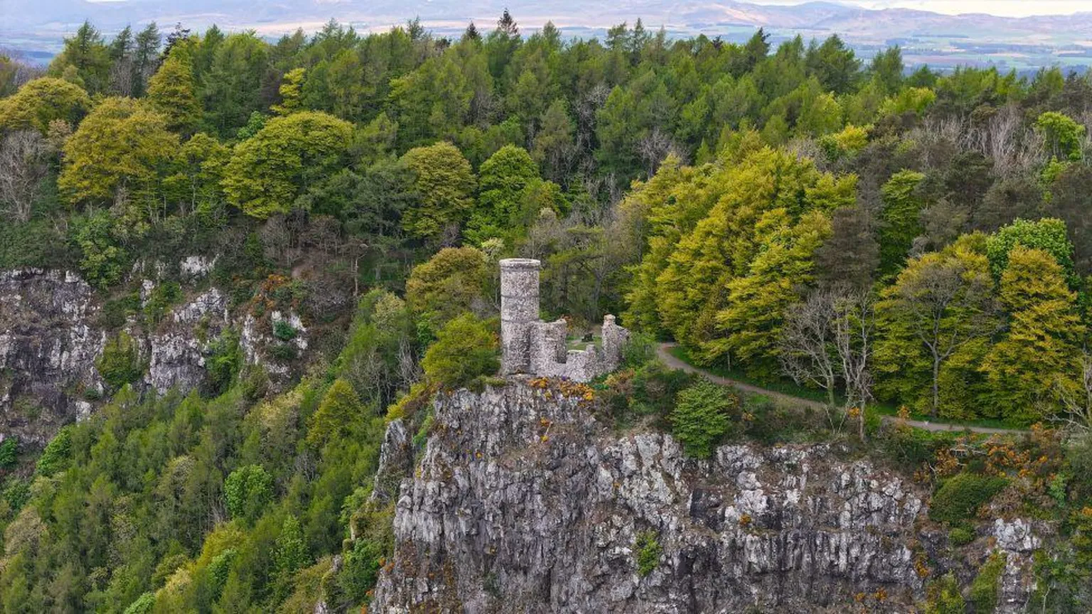 Kevin Hamill An old tower nestled on the side of a hillside, in among trees, with hills in the far distance.