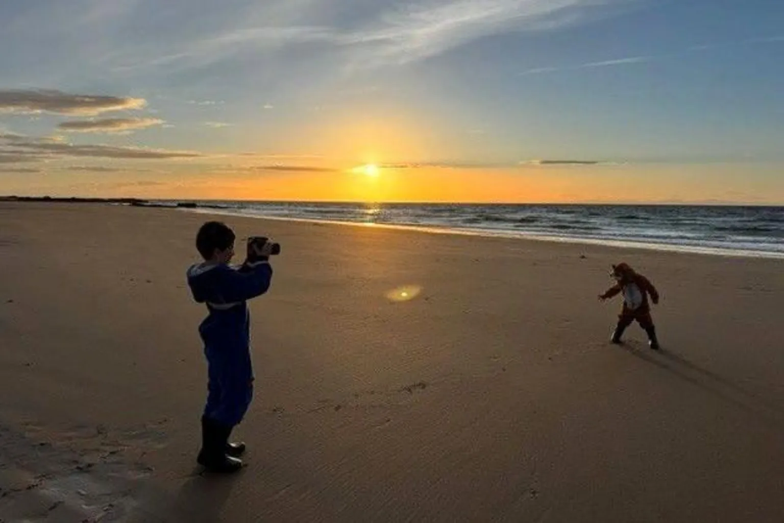 Zoe Mann Two young boys on a beach at sunset, one is holding a camera, the other is making a dance shape, the sun is glowing in the distance over the sea.