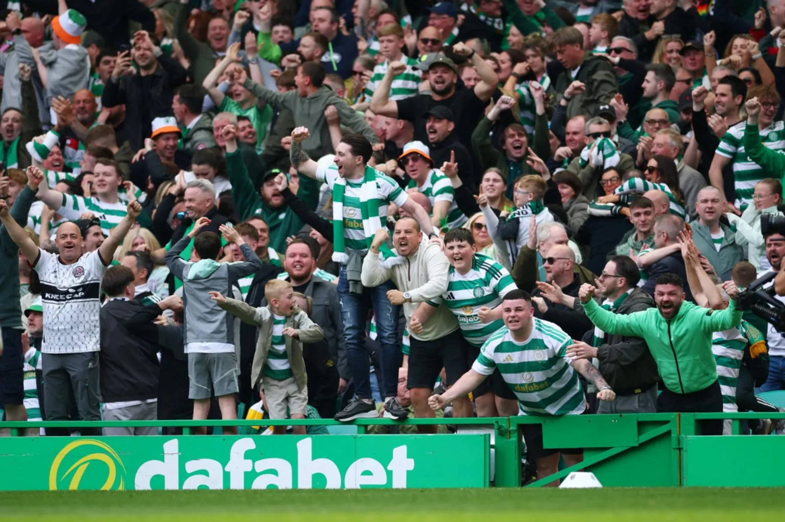  A packed crowd of Celtic supporters celebrate wildly in the stands during a Scottish Premiership match at Celtic Park. Fans wearing green-and-white striped shirts and scarves cheer, raise their fists, and embrace as excitement spreads through the stadium.