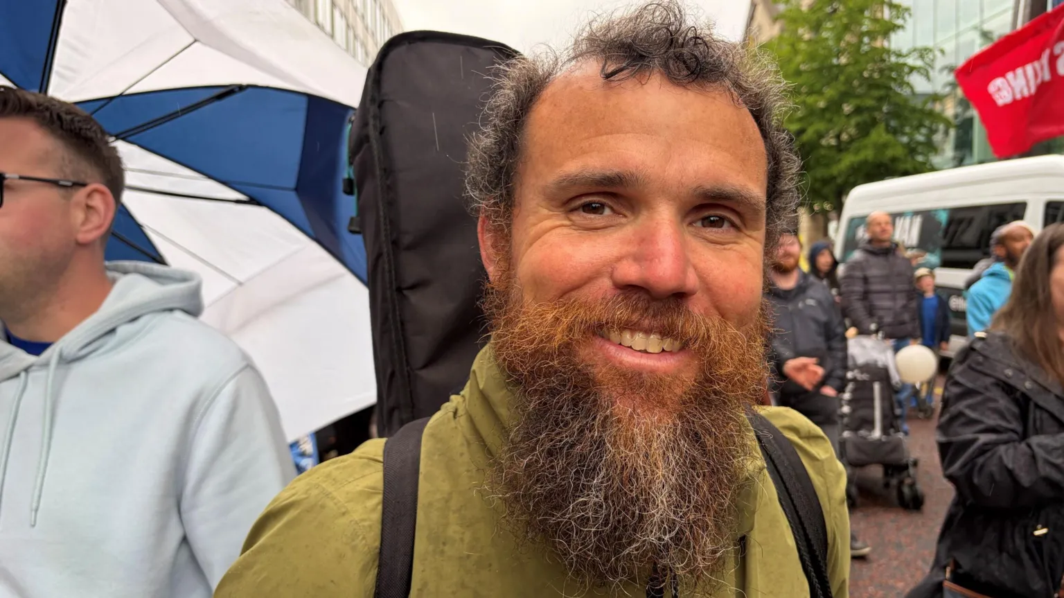 A man with short-brown hair and a long brown beard smiles to the camera. Behind him is a crowd of people, out-of-focus. The man wears a green waterproof jacket and has a guitar case on his back.