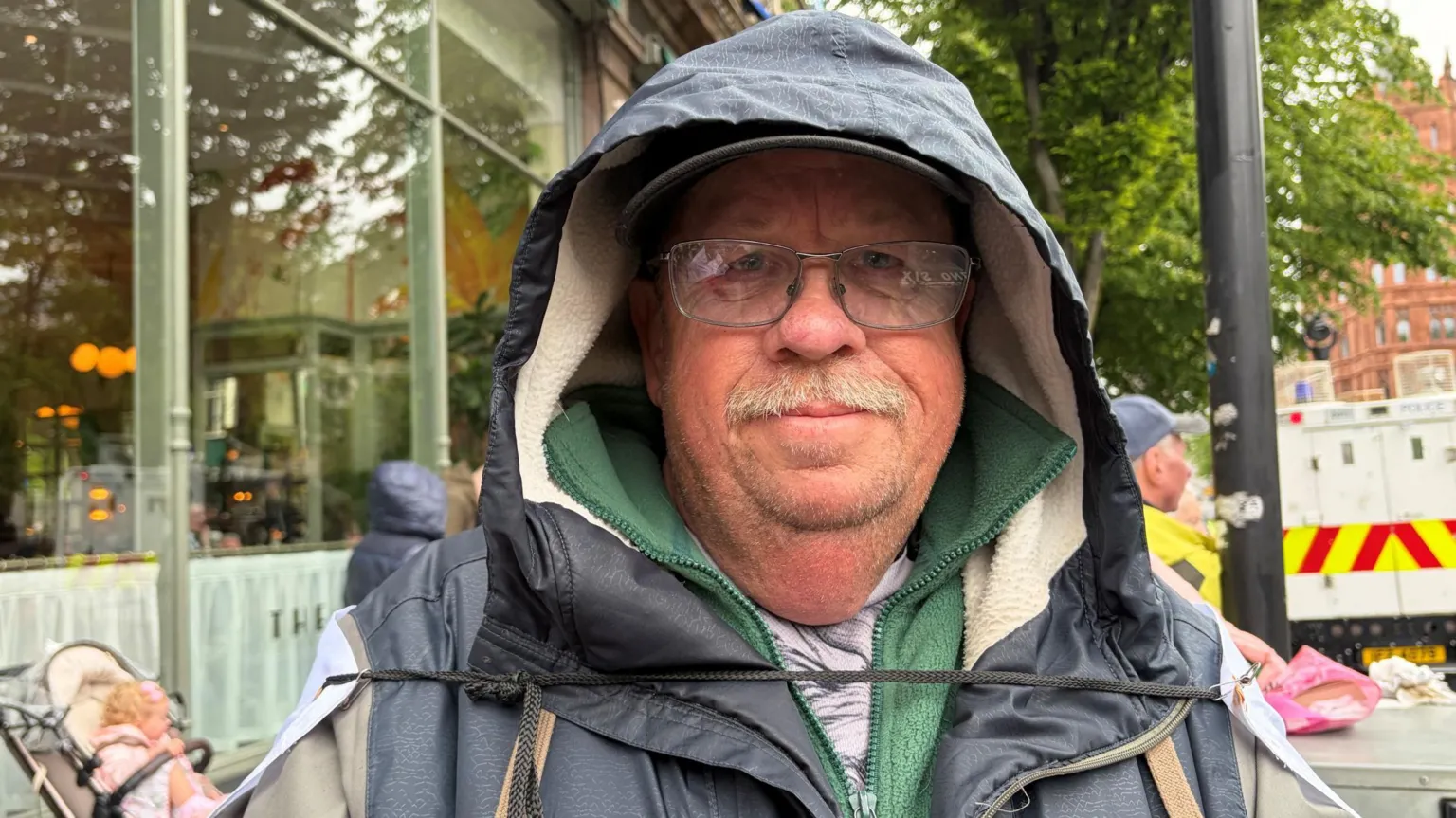 A man with clear spectacles and a grey moustache stairs to the camera. He wears a navy waterproof jacket, with a green fleece underneath. Over his left shoulder is a white police van. Over his right, a baby sits in a cream pram below a clear window.
