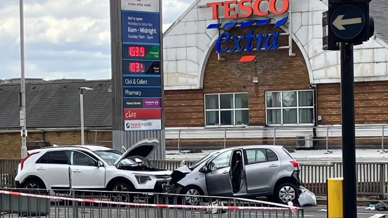A Tesco Extra can be seen in the background of the shot along with the two collided vehicles before a metal fence. A police cordon is closing off the area.