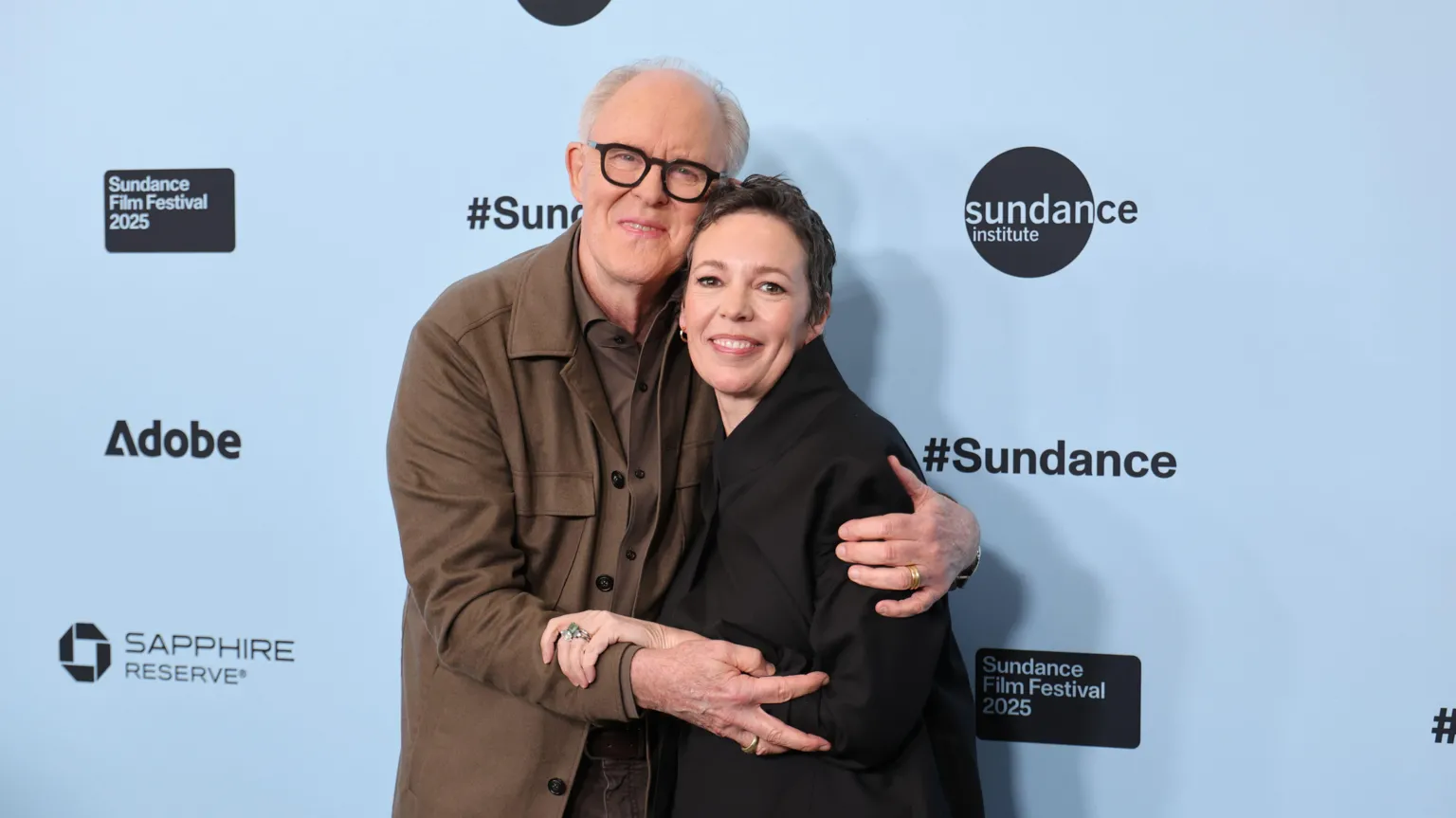 Neilson Barnard / John Lithgow iswaring a brown shirt and top and hugging Olivia Colman who is in a black jacket. They attend the