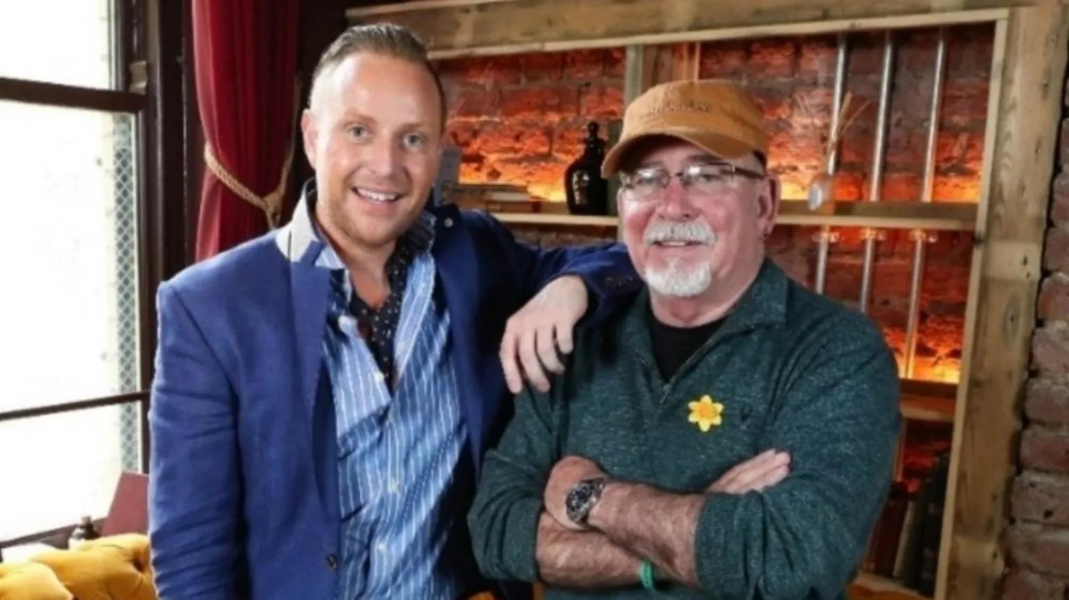 Marcus Hunter-Neill Marcus (left) has his arm resting on the shoulder of Billy. Marcus is wearing a blue suit jacket and a white and blue striped shirt with a dark blue neck scarf. He has short slicked back ginger hair. Billy has his arms crossed. He is wearing a dark green half zip top and has a daffodil badge on the front. He is wearing a brown flat cap. 