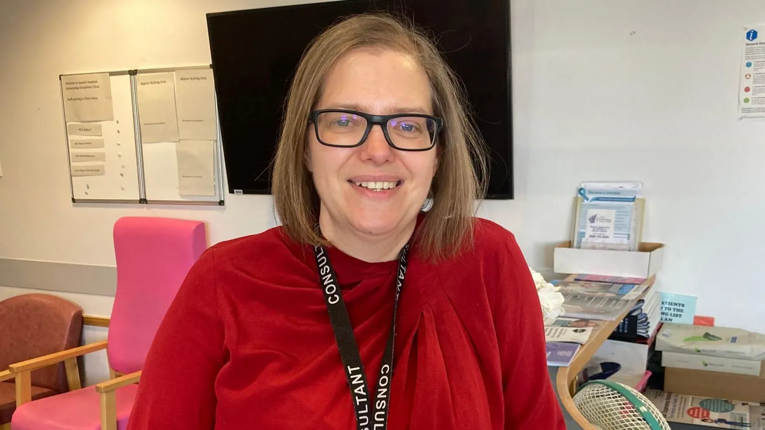 East Suffolk and North Essex NHS Foundation Trust A woman with light brown hair in a bob style sits in a chair within a hospital office. She is smiling. She wears glasses, a red blouse and a black lanyard around her neck.