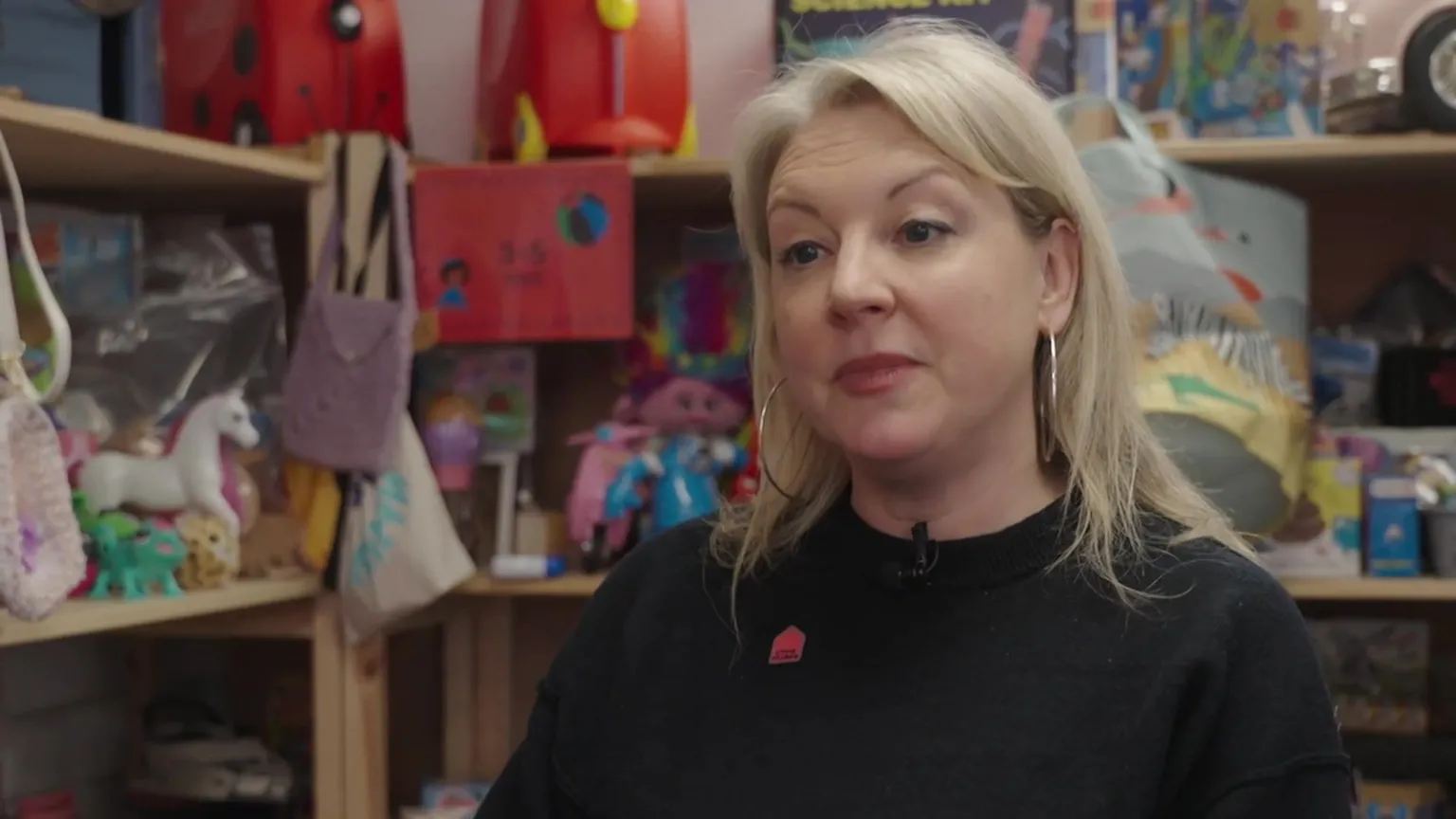 Sophie Livingstone seated indoors in front of shelves filled with toys, books and colourful items, wearing a dark top with a small badge, in a close interview shot.