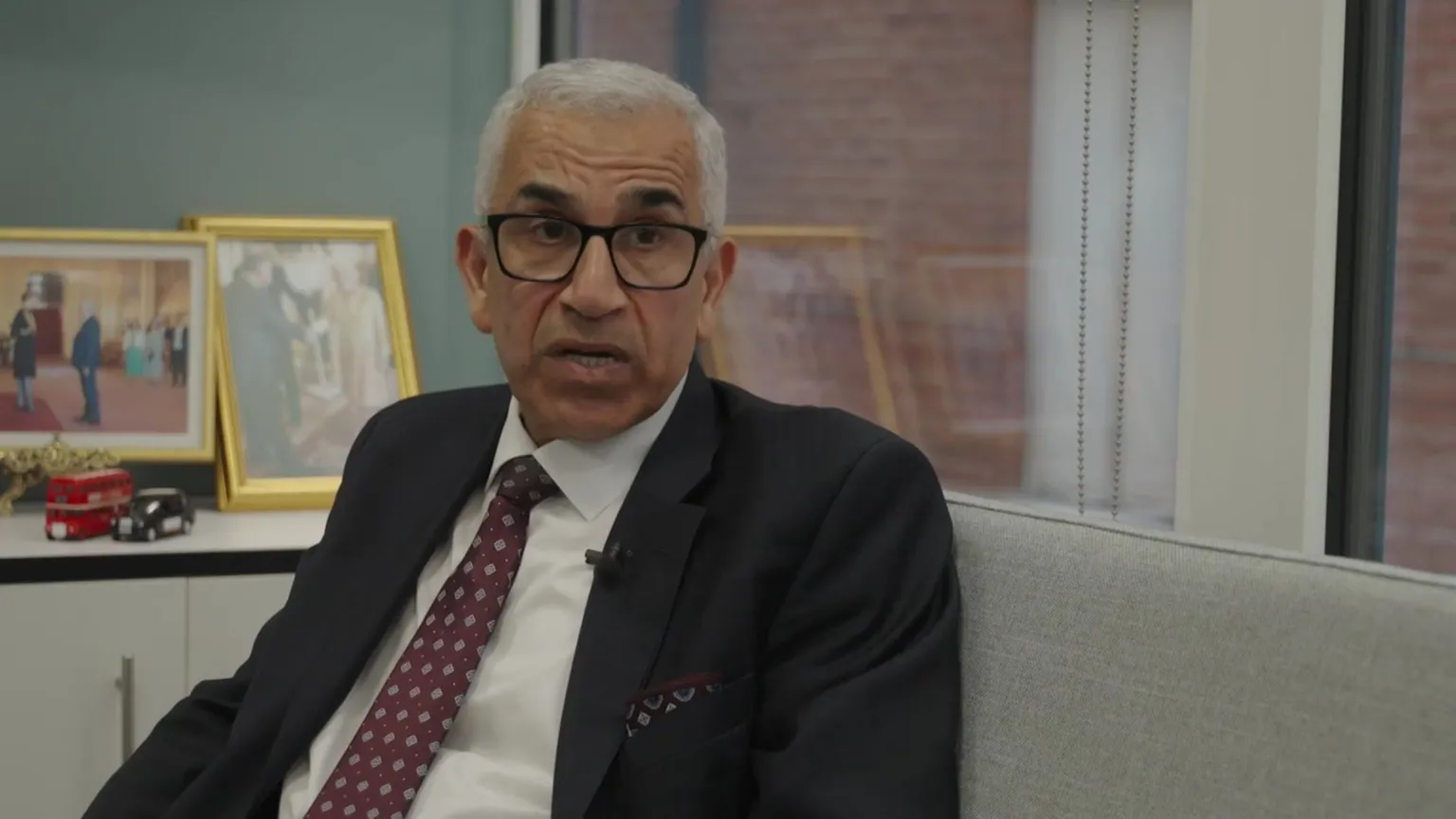 Person in a suit and patterned tie seated on a sofa in an office setting, with framed photographs and model buses visible on a shelf behind, during an interview.