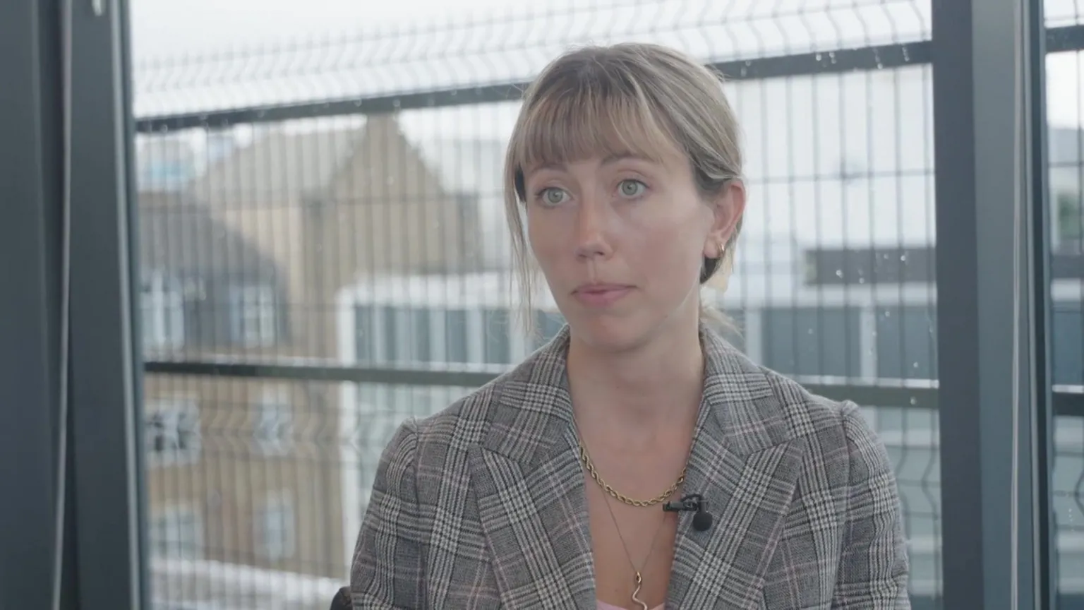 Antonia Jennings wearing a checked blazer and necklace, seated indoors in front of a large window with metal railings visible outside, speaking during an interview.