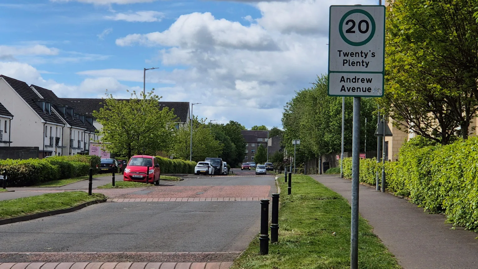A view looking down a street, showing rows of houses on either side and various parked cars. A sign reading Twenty's Plenty - Andrew Avenue is at the top of the street.