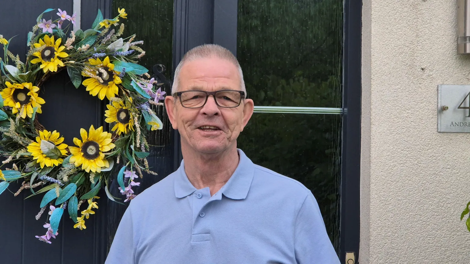 A man standing in front of his house and smiling for the camera. e has short white hair, glasses and a blue top on.