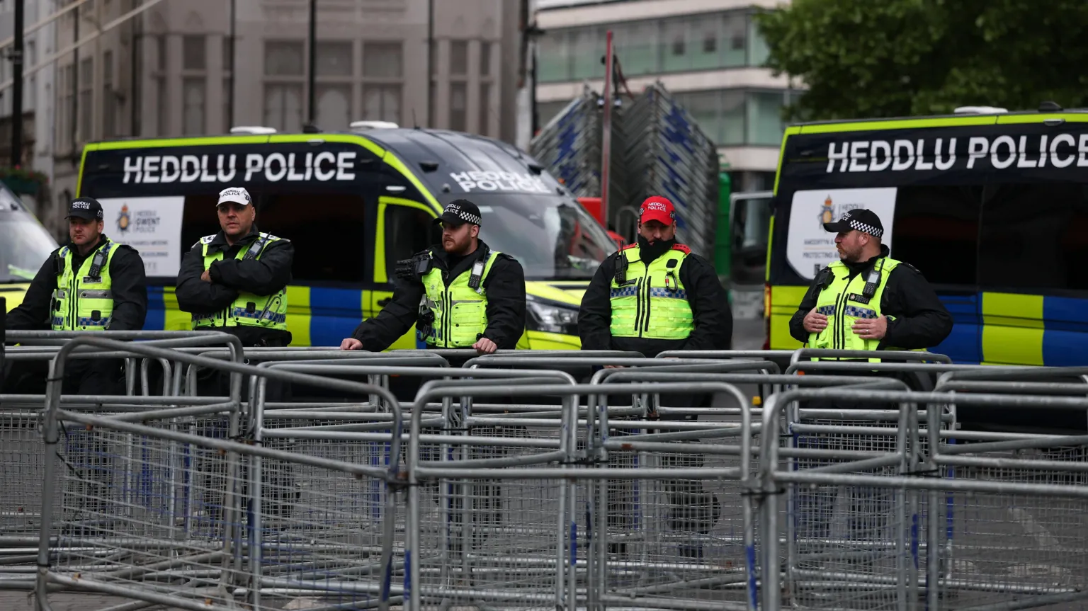  Police officers stand behind a row of metal barriers in front of two police vans which say HEDDLU POLICE in dual Welsh and English signage.