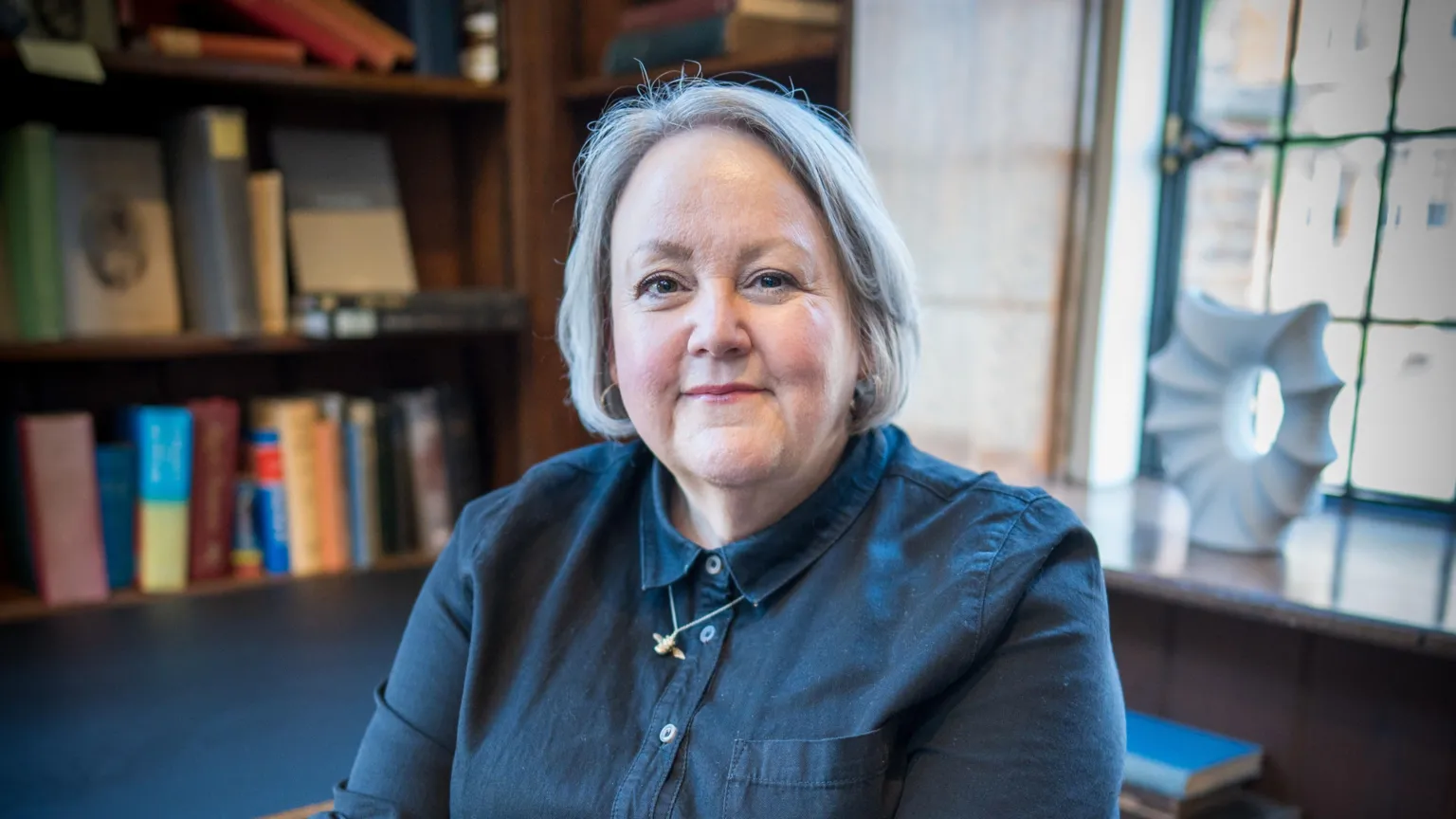 Claire Malcolm, a woman with grey hair and a blue shirt sat in front of a book case.