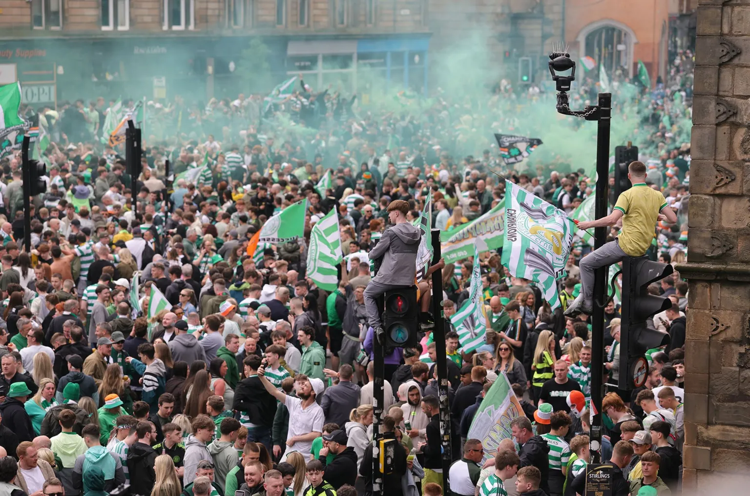 A large crowd of Celtic supporters gathers in a Glasgow street, with green smoke in the air and flags being waved, while some people sit on traffic lights.