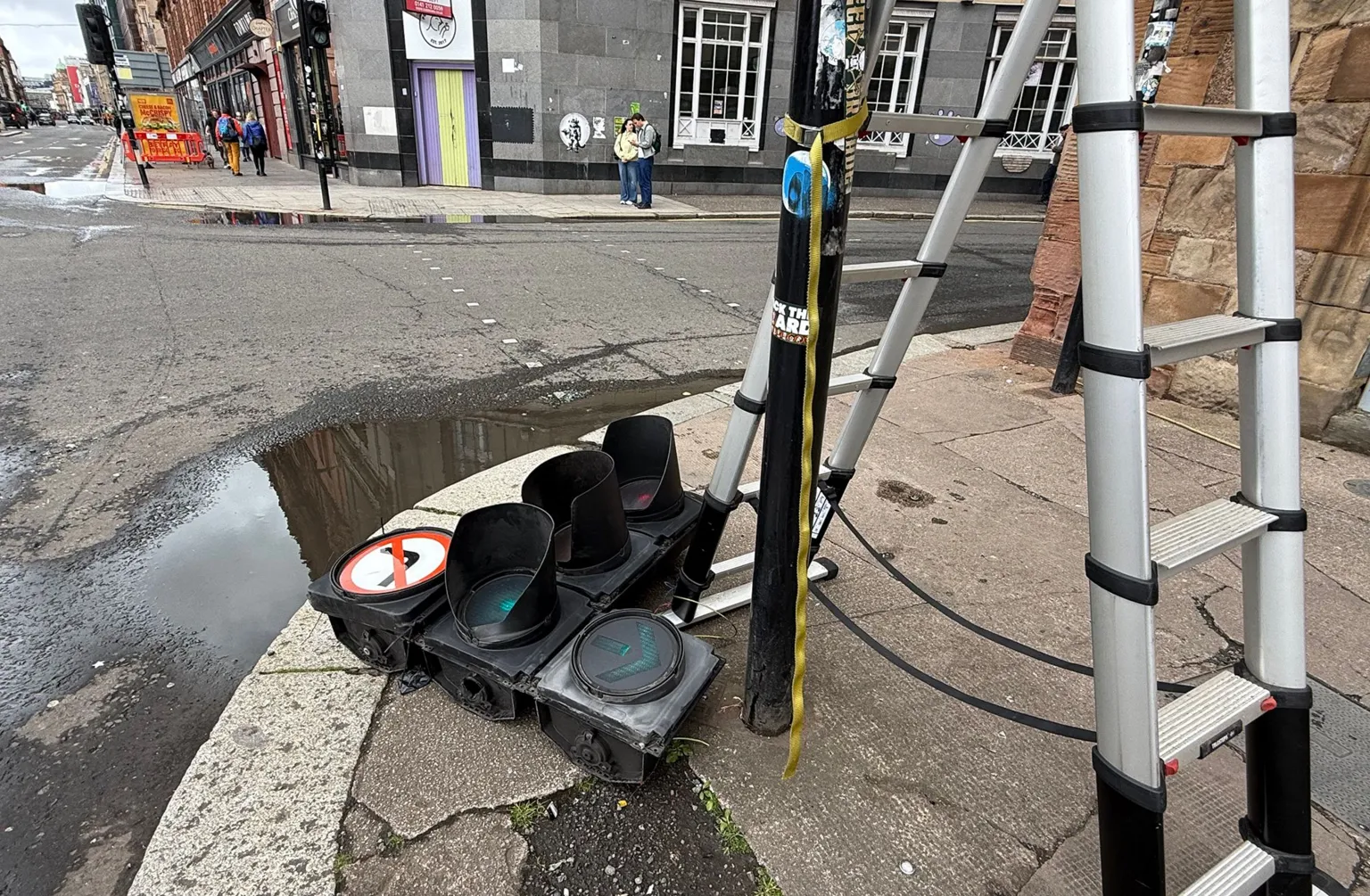 A damaged set of traffic lights lies on the pavement beside a road in Glasgow, with a ladder strapped to a nearby pole and debris visible.