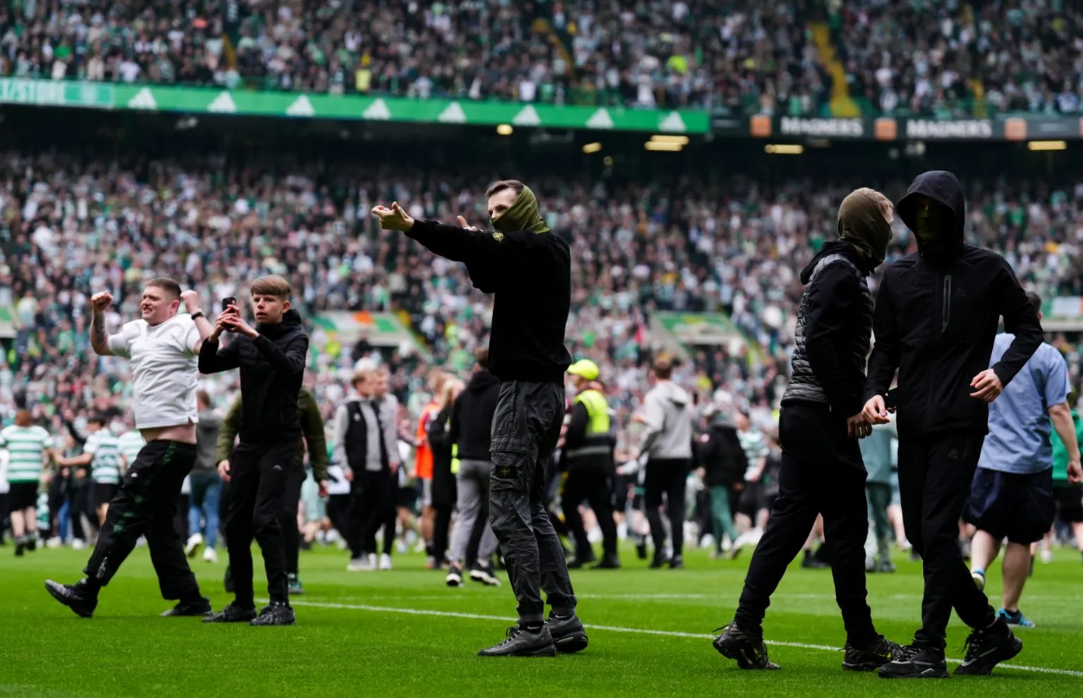 Several people stand on the pitch at a crowded football stadium, with more supporters in the stands behind them after fans spilled onto the field.