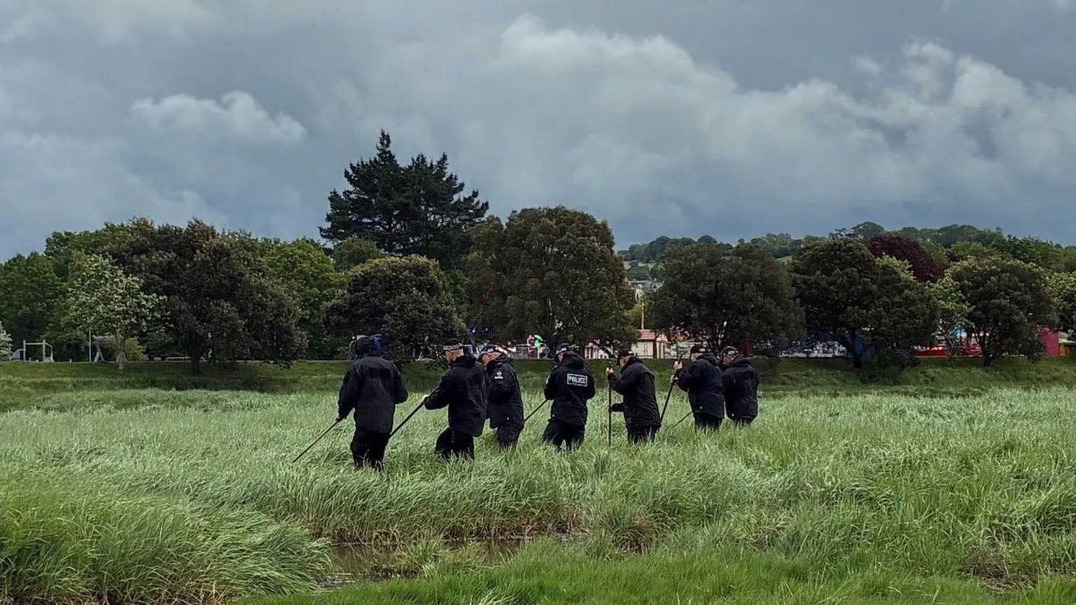 Devon and Cornwall Police Seven police officers are seen in the middle of a field of tall grass with a body of water to the left and trees in the background beneath a moody cloudy sky. 