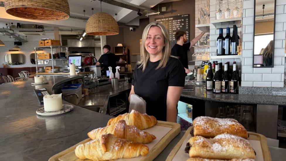 Llun o Nia Roberts wrth gownter ei bwyty - Baravin - gyda croissants ar y cownter ac aelodau staff yn parhau a'u gwaith yn y cefndir