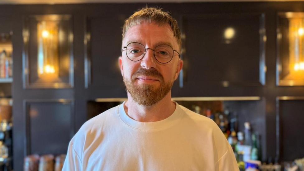 Llun o Gareth Evans - dyn sydd a barf brown a sbectol crwn, mae'n gwisgo crys T gwyn ac mae'n sefyll o flaen bar Gwesty Cymru 