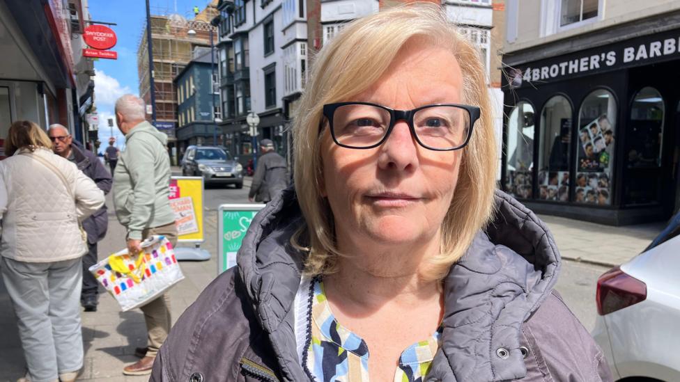 Llun o Lynn Phillips yn sefyll yng nghanol y dref yn Aberystwyth - sbectol a chot lliw piws