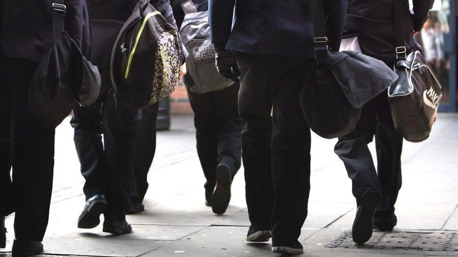 Group of students in school uniforms walking along a pavement, each carrying shoulder bags and backpacks