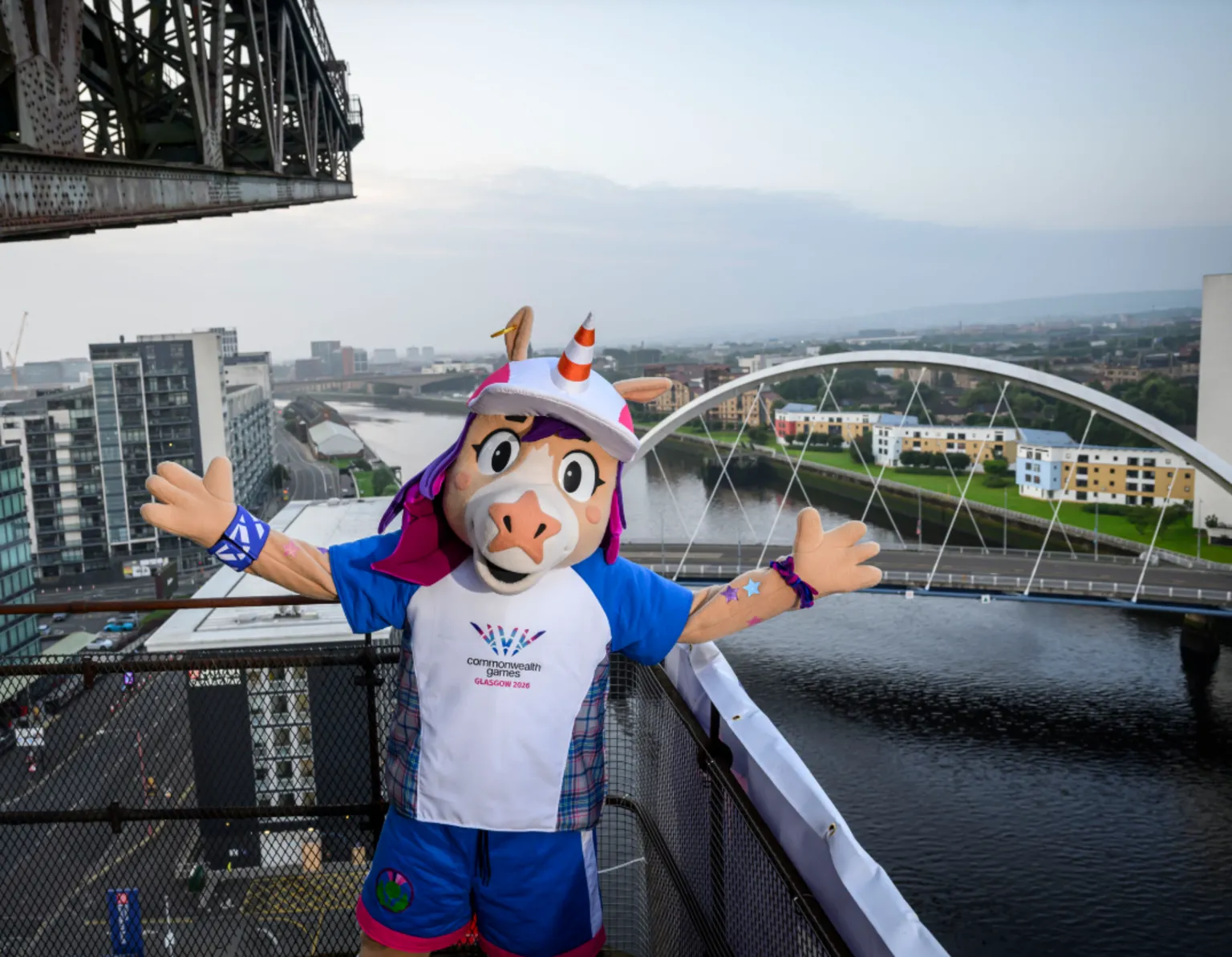 Glasgow 2026 The Glasgow 2026 Commonwealth Games mascot stands on a platform overlooking the River Clyde, with the white arch of the Clyde Arc bridge and city buildings in the background, while part of a large crane structure is visible overhead.