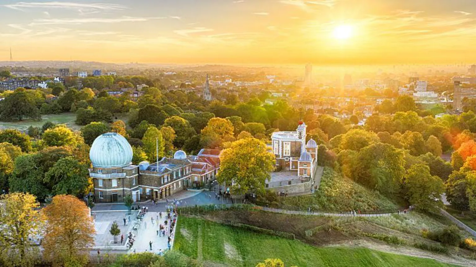 The Royal Observatory as seen from the sky. It has a large domed building on one side and a plaza at the front. In the background is the London skyline and the setting sun.