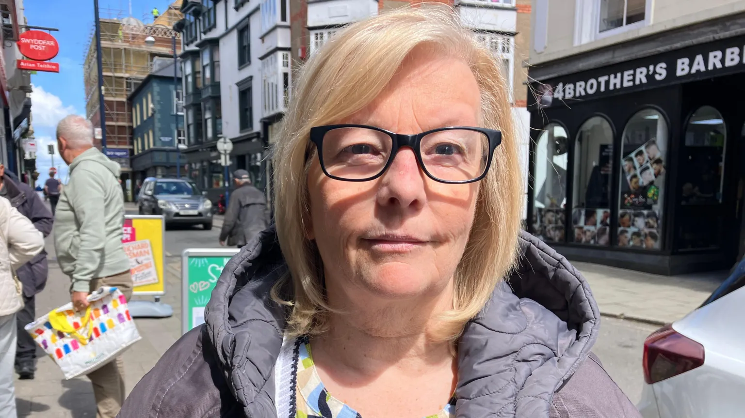 A photo of Lynn Phillips - wearing glasses and a lilac coloured coat standing in Aberystwyth town centre 