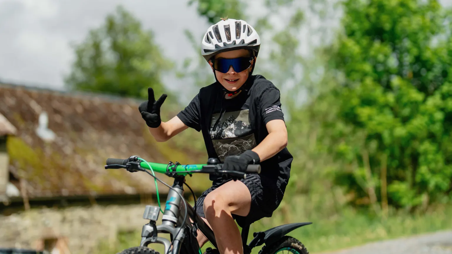 Red On Sports A young boy on a bike holds up two fingers for victory while pedalling a green bike. He is in a black top and shorts with a white helmet on and dark glasses.