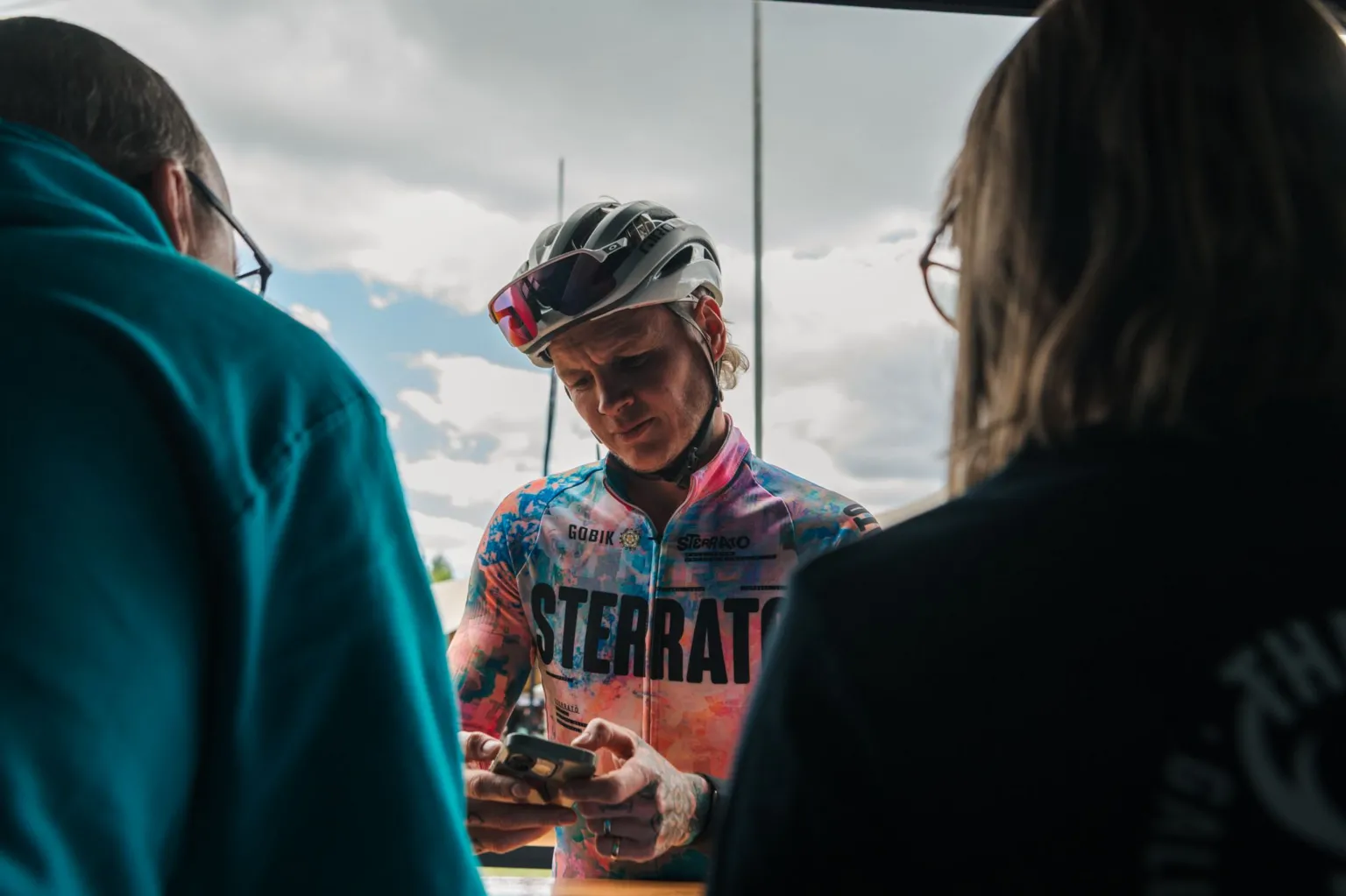 Red On Sports A cyclist in a brightly coloured top with their sunglasses up on top of their helmet pauses to take a picture on his phone. He is viewed from between two people looking on that we can only see in outline.