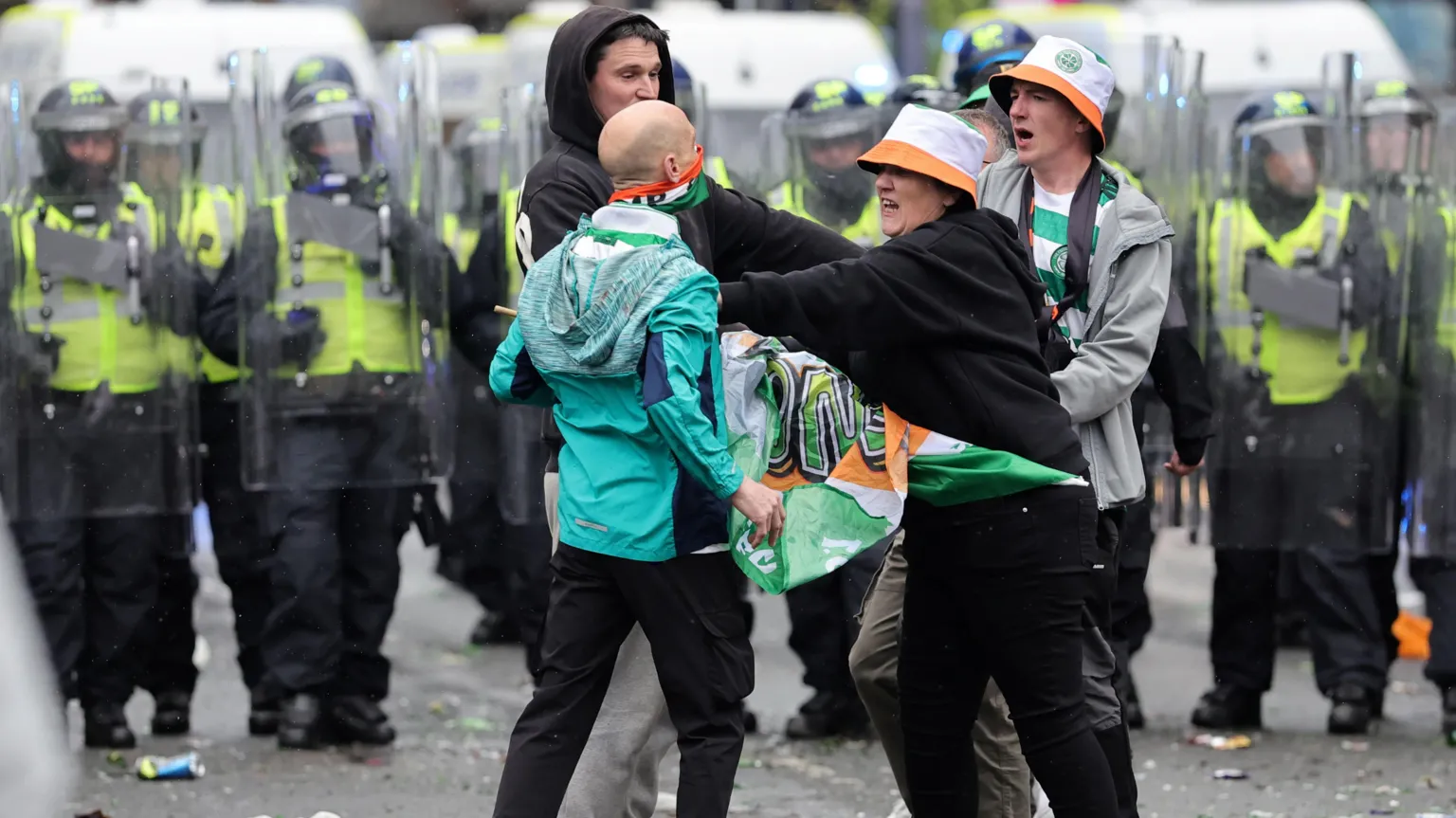  A Celtic fan dressed in black and wearing an Irish tricolour bucket hat holds back a fellow supporter in a green jacket as a two men look on in a city street. Officers holding riot shields can be seen in the background.
